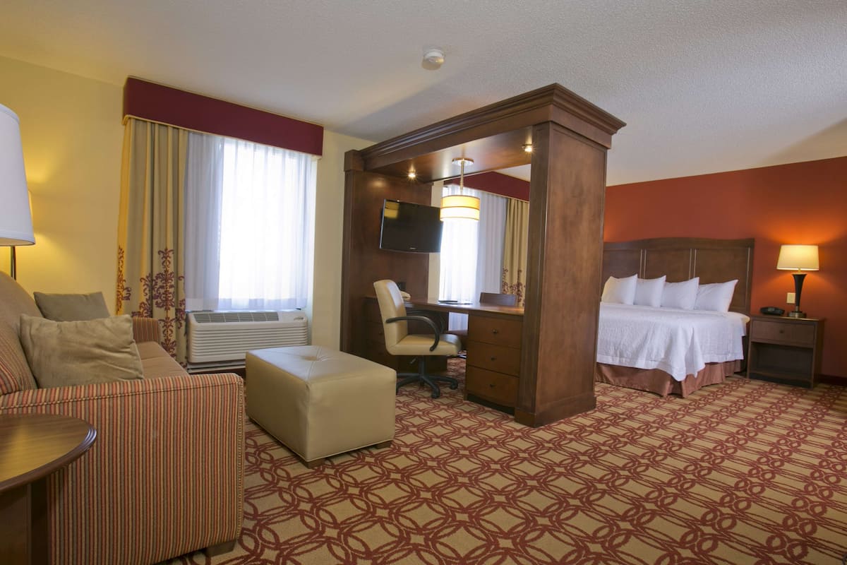 A hotel room with a bed, a desk, a chair, an air conditioning unit, and a television.