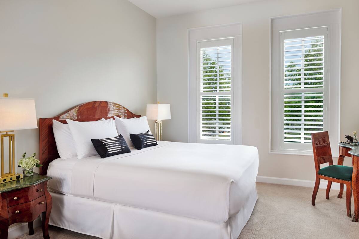 A neatly arranged bedroom with a large bed, two bedside tables with lamps, a chair, and a window with blinds.