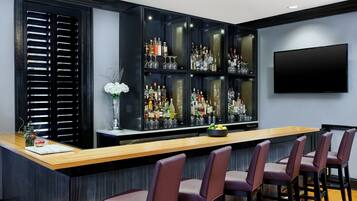 Zona de bar con mostrador de madera, varias estanterías llenas de botellas y cuatro taburetes.