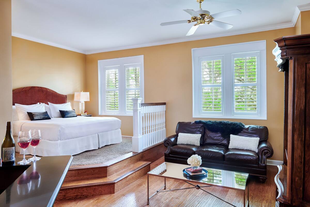 A hotel room with a bed, a leather sofa, a coffee table, and a desk with a bottle and glasses.