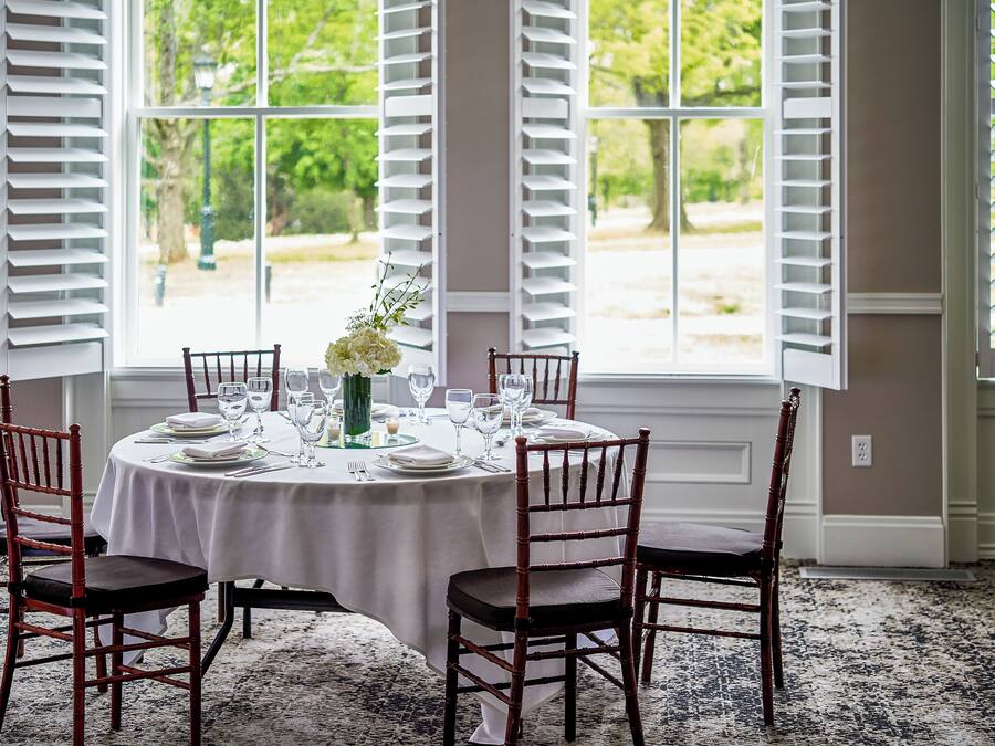 A round table set for six with white tablecloths, wooden chairs, and a vase of flowers.