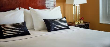 Habitación de hotel con una cama grande, almohadas blancas y negras, cabecera de madera, mesita de noche con lámpara y una ventana con cortinas.