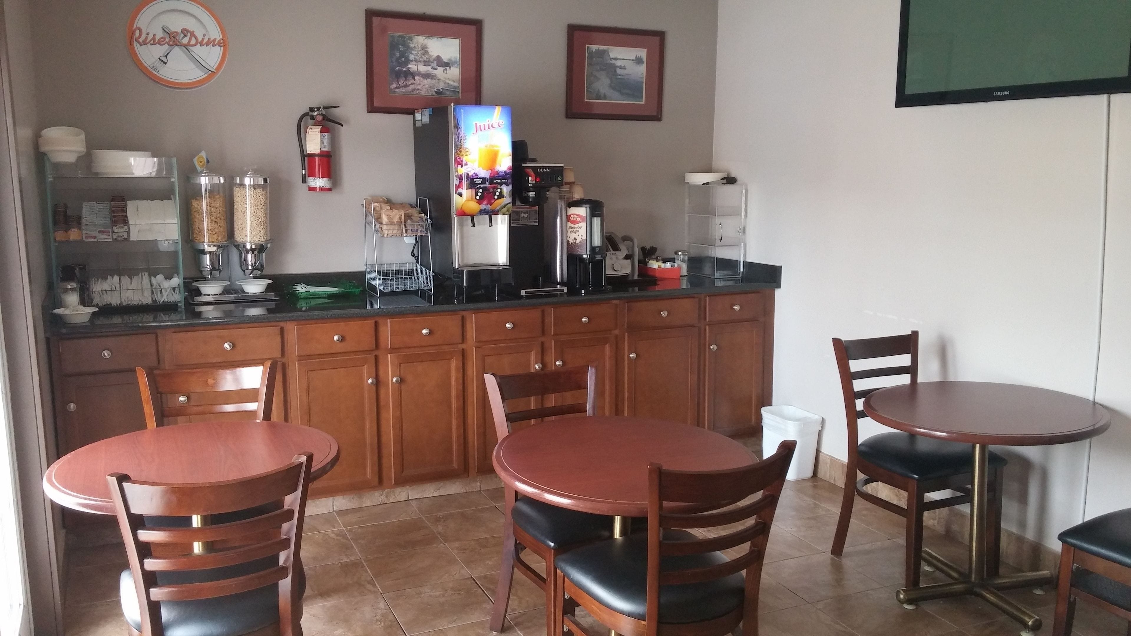 A small, clean kitchen with wooden cabinets, a coffee machine, and a juice dispenser. There are two wooden tables with chairs, a clock, and framed pictures on the wall.