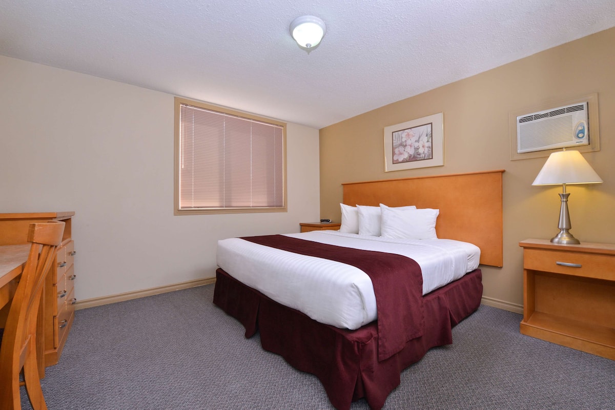 A hotel room with a large bed, a desk, a chair, a window with blinds, a bedside lamp, and an air conditioning unit.