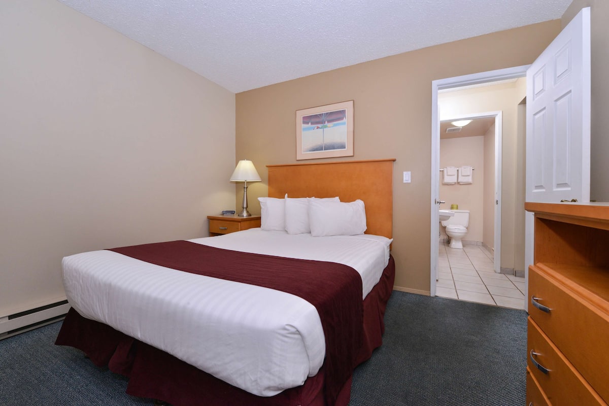 A hotel room with a large bed, a nightstand with a lamp, a wooden dresser, and a bathroom visible through an open door.