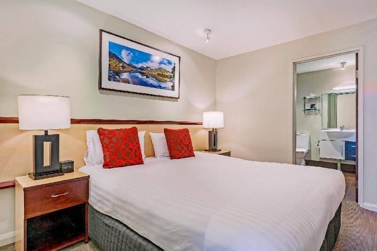 Habitación de hotel con una cama grande, dos lámparas de noche, una mesita de noche con cajón, un cuadro en la pared y un baño visible a través de una puerta abierta.