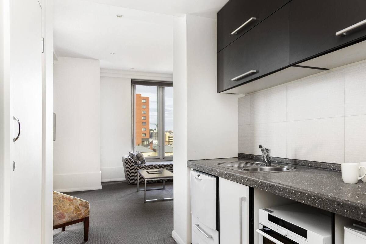 Una cocina moderna con un lavamanos, encimera y armarios. Hay una zona de comedor con una silla y un sofá. Una ventana ofrece vistas de edificios.