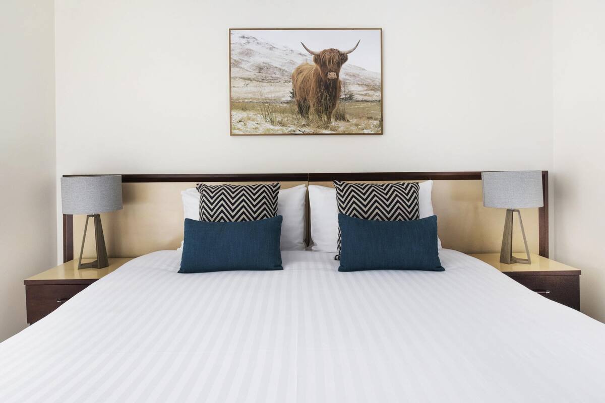 Habitación de hotel con una cama, dos mesitas de noche, dos lámparas y un cuadro de un toro en la pared.