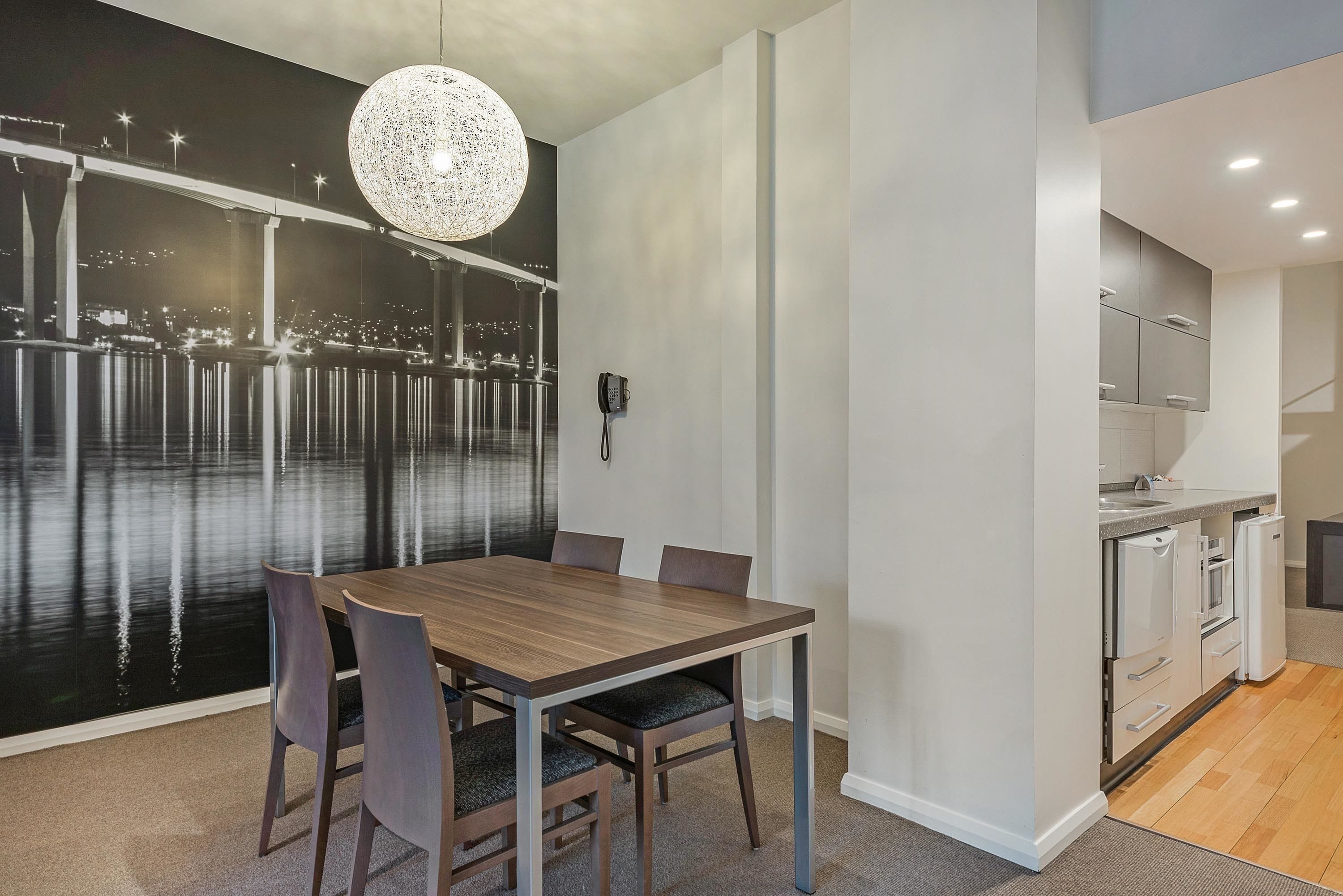 A modern dining area with a large table and chairs, a decorative wall with a cityscape, and a kitchen with white appliances.