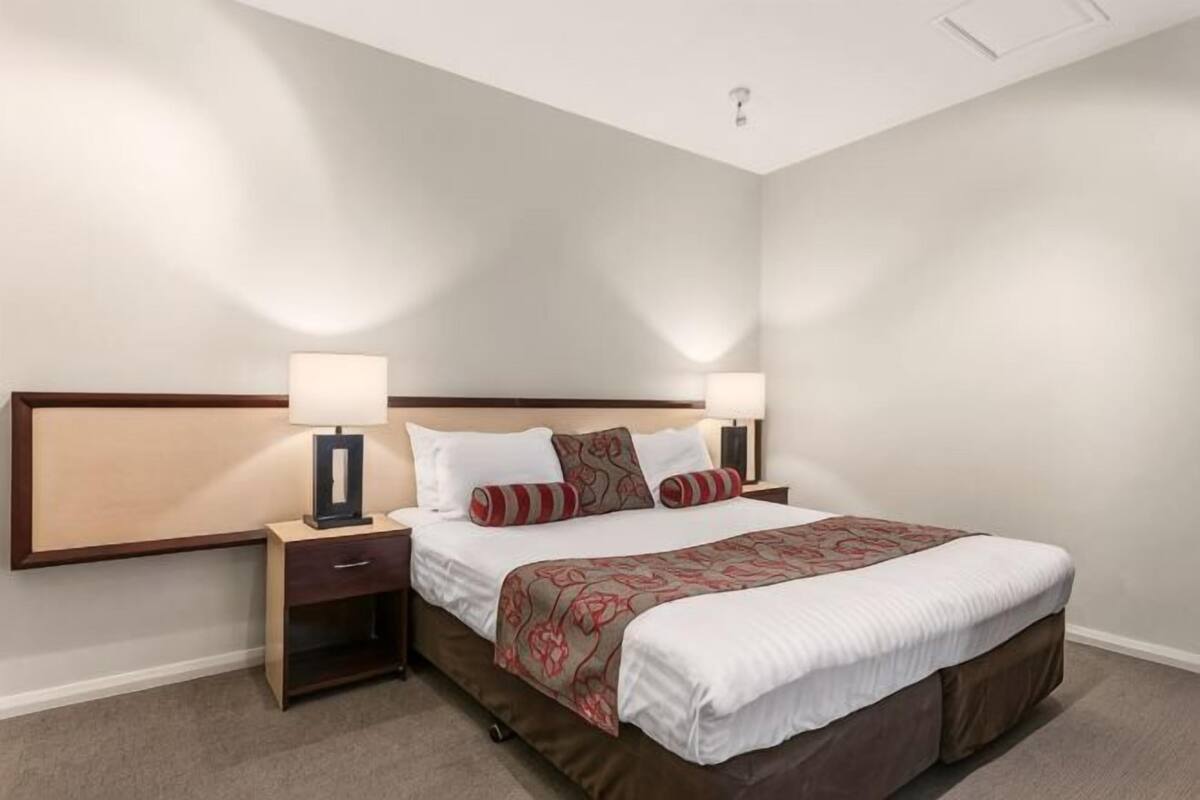 Una habitación de hotel con una cama, dos mesitas de noche con lámparas, un cabecero y piso alfombrado.