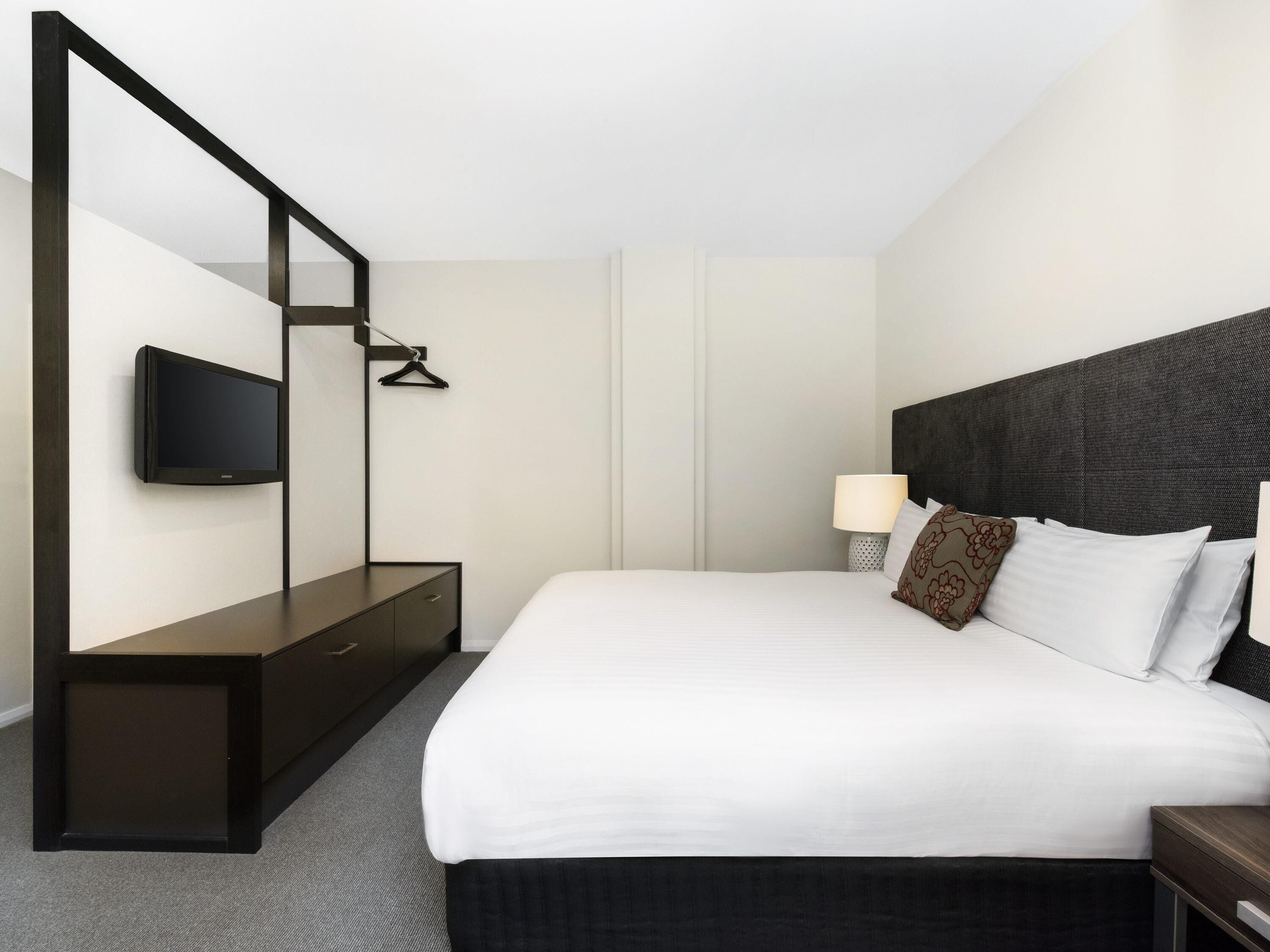 A hotel room with a bed, a TV mounted on the wall, a bedside table, and a lamp.