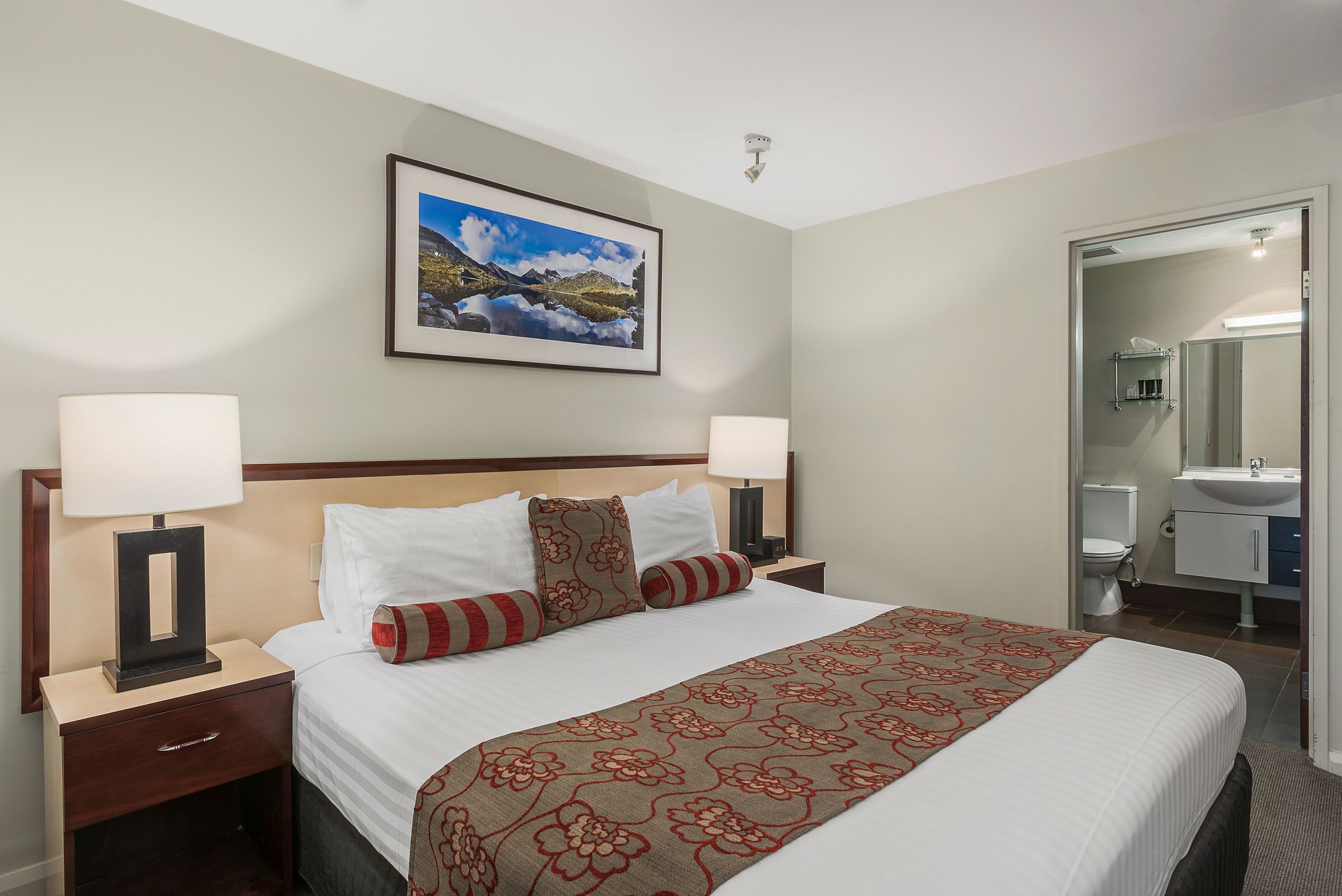 A hotel room with a large bed, two bedside lamps, a framed landscape picture, a bathroom visible through an open door, and a nightstand with a drawer.