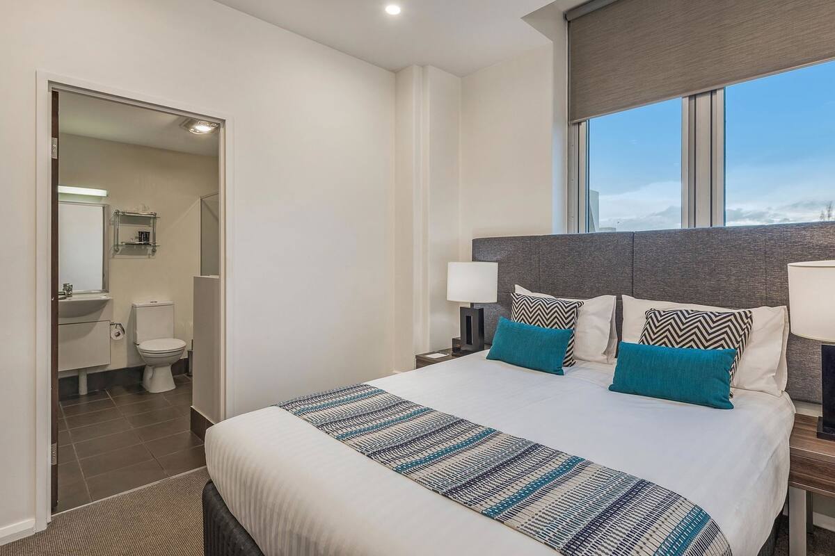 Habitación de hotel con una cama grande, una mesita de noche con lámpara, vista al cielo y baño visible a través de una puerta abierta.