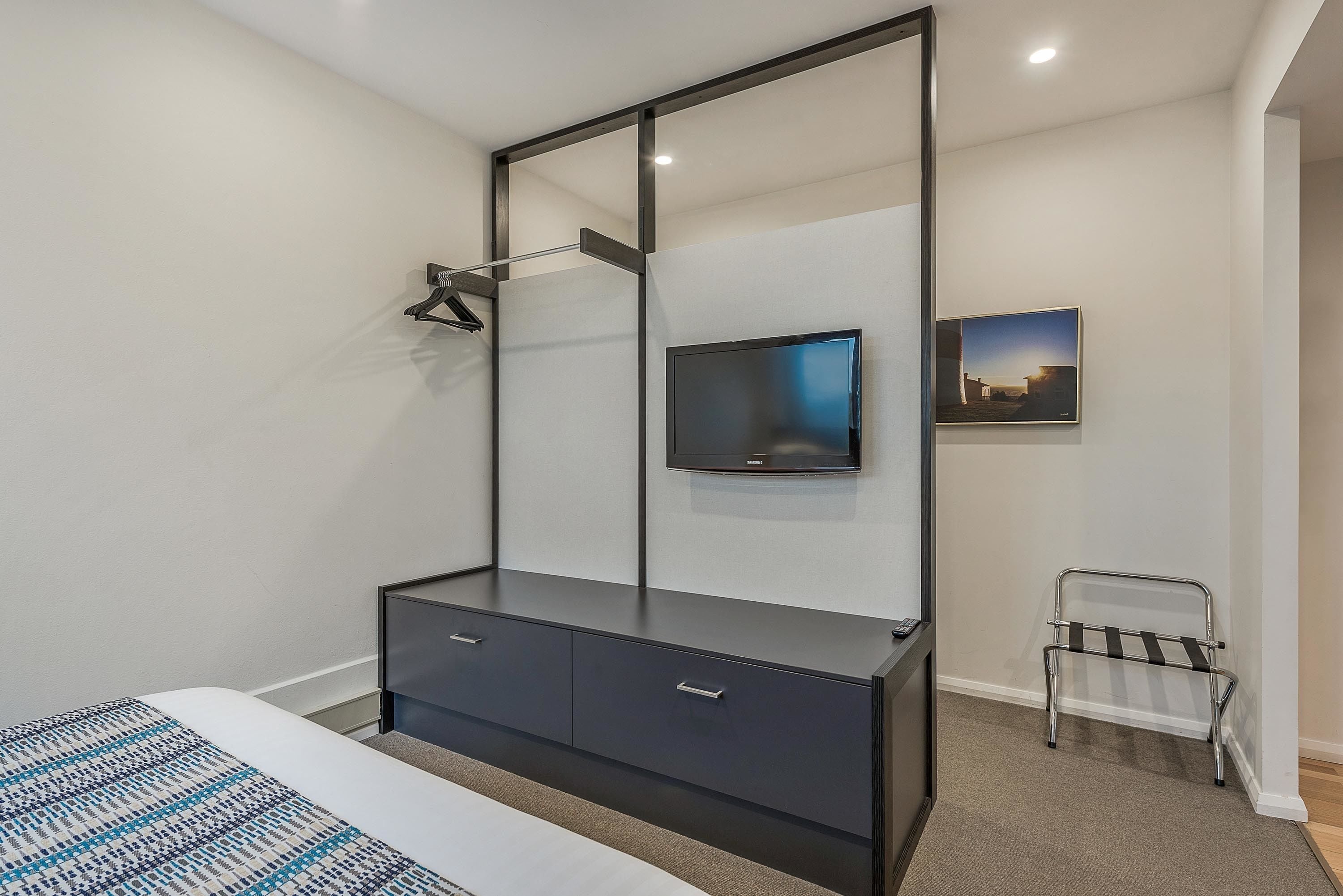 A modern bedroom with a large bed, a dark dresser with drawers, a mounted TV, and a small seating area with a chair and a bench.