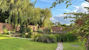Um jardim tranquilo com lago, vegetação exuberante e um muro de tijolos com janelas em arco.
