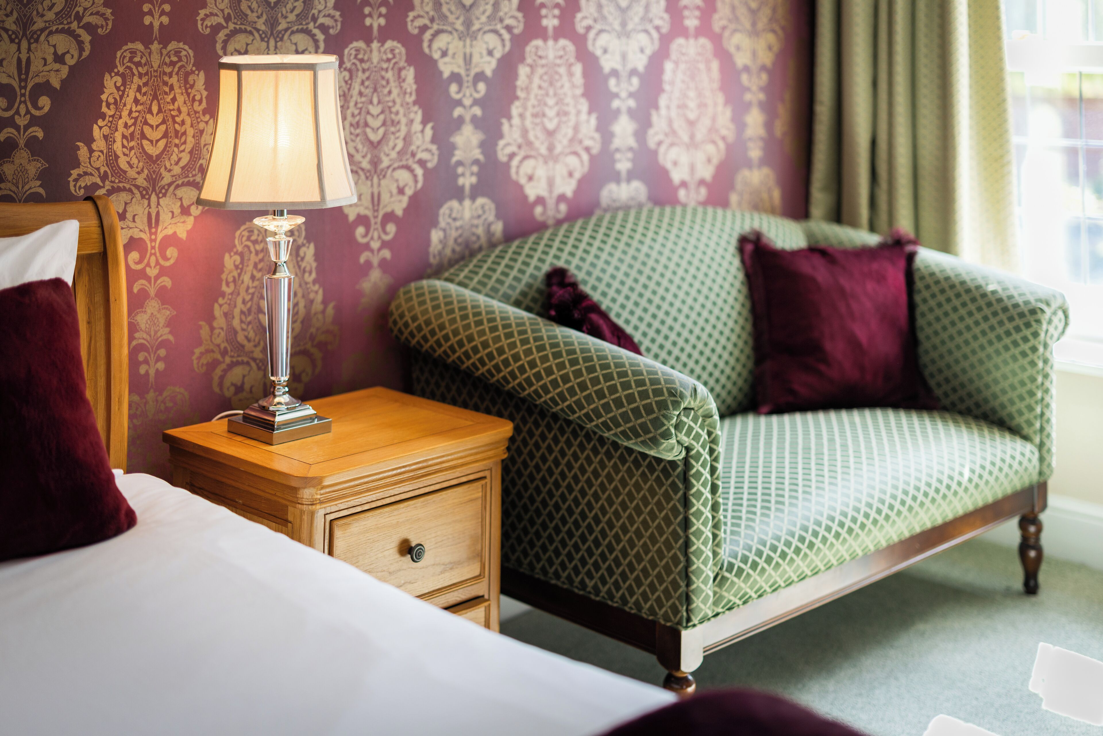 A hotel room with a green patterned sofa, a wooden nightstand with a lamp, and a bed with white linens.