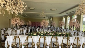 Uma sala de banquetes com mesas redondas, toalhas brancas e cadeiras adornadas com cortinas brancas.