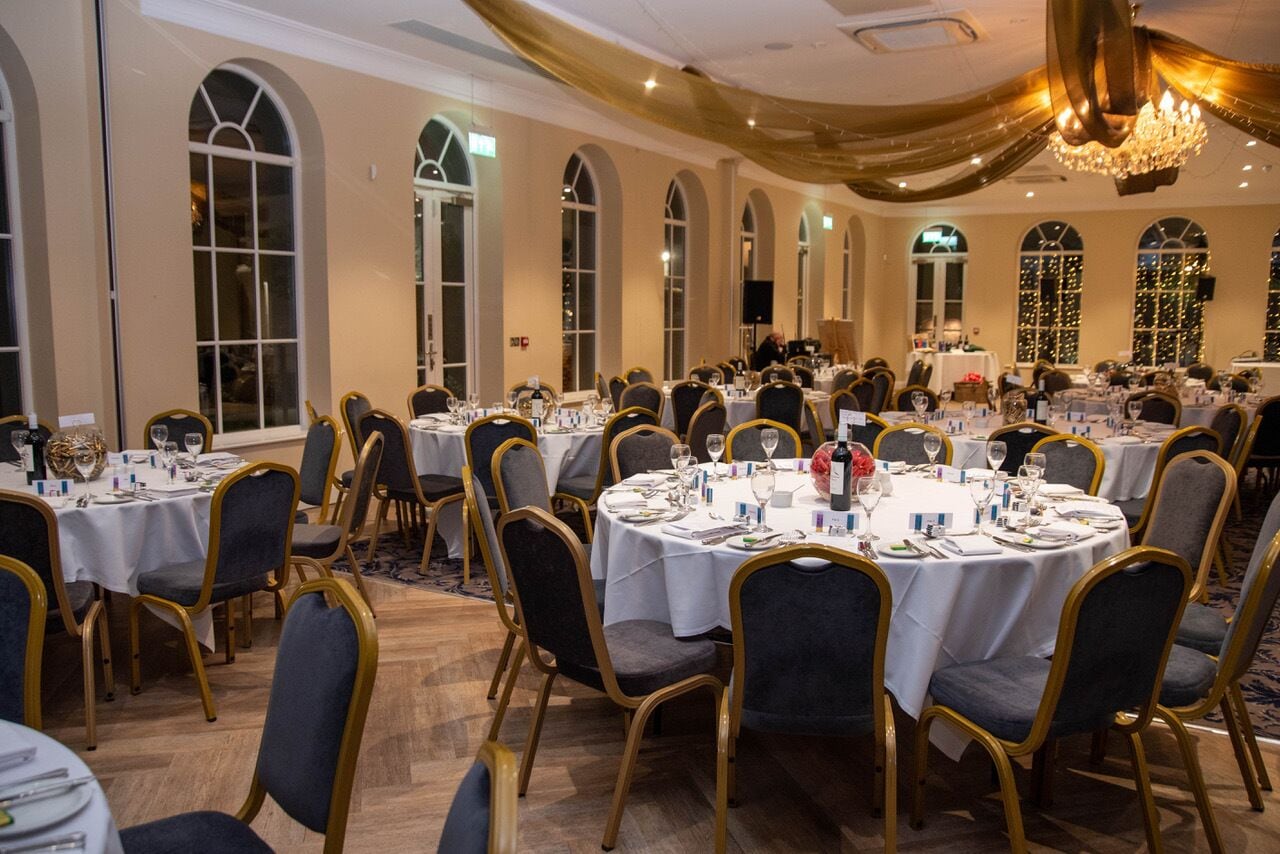 A banquet hall with round tables, gold chairs, and chandeliers.