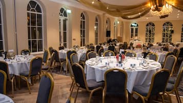 A banquet hall with round tables, gold chairs, and chandeliers.