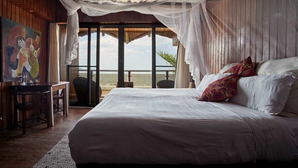 A bedroom with a large bed, a desk, and a painting. There is a view of the ocean through large windows. - Catembe Gallery Hotel (Maputo)