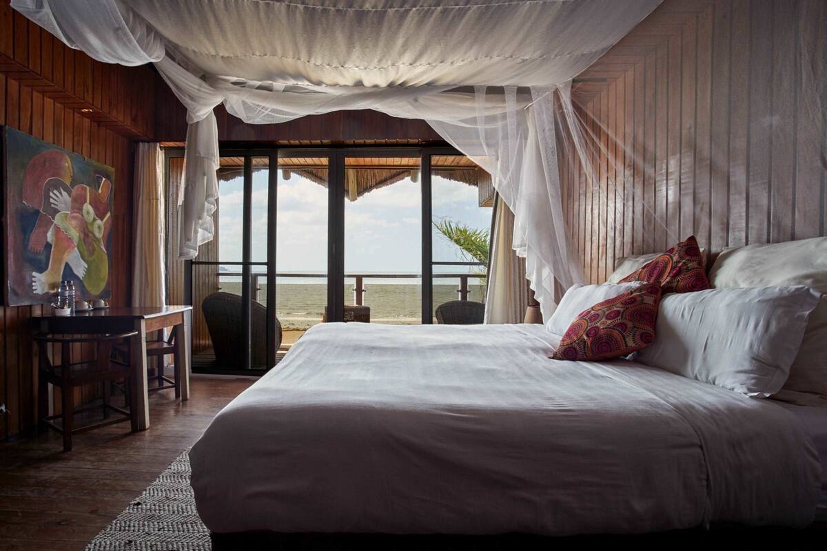 A bedroom with a large bed, a desk, and a painting. There is a view of the ocean through large windows.