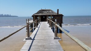 A wooden pier leading to a small building with a thatched roof, situated on a beach with a city skyline in the background. - Catembe Gallery Hotel (Maputo)