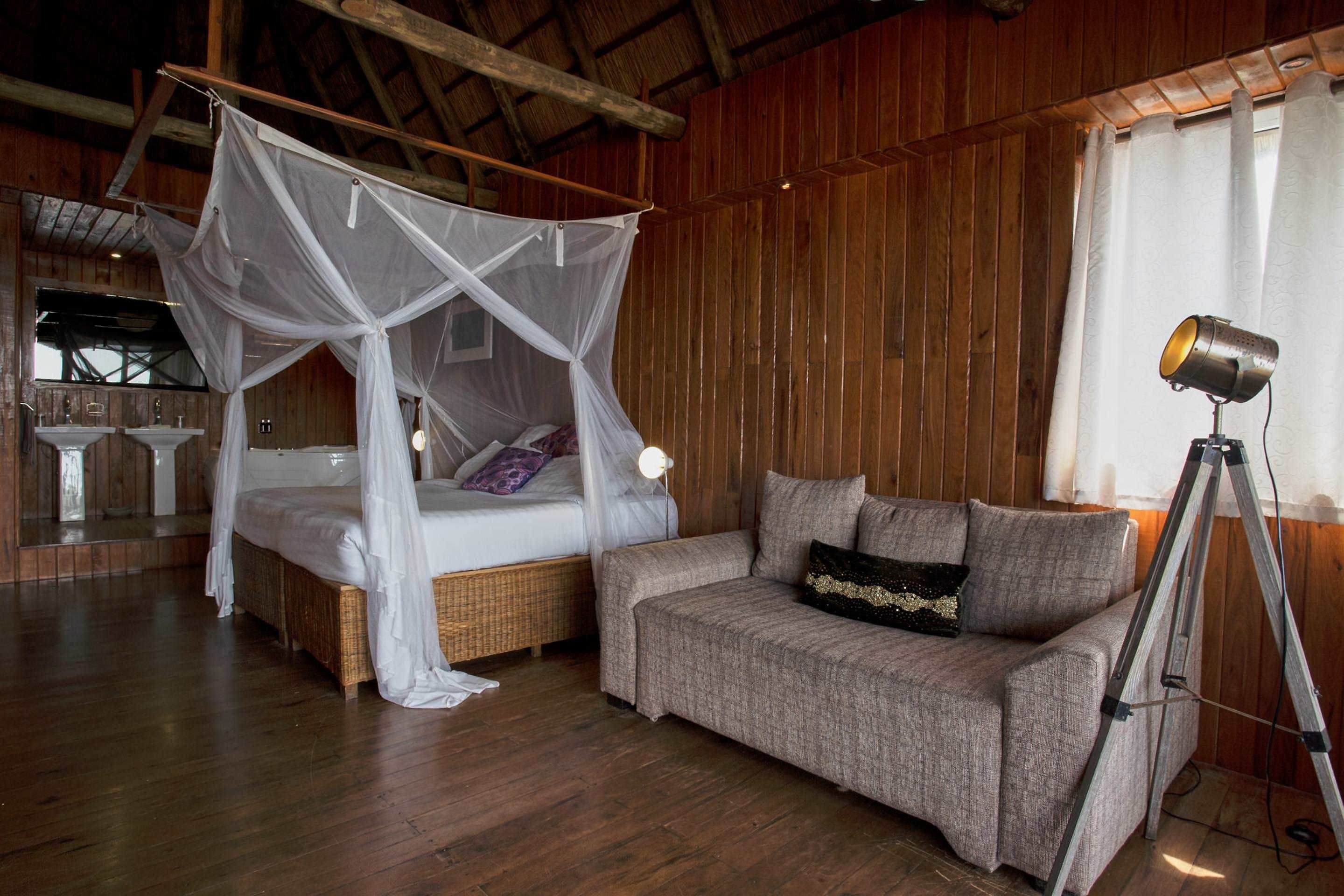 A wooden room with a bed under a canopy, a sofa, and a tripod with a lamp.