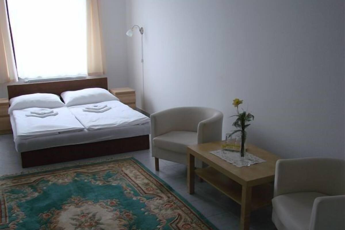 A hotel room with a bed, two chairs, a small table with a vase, a floor lamp, and a window with curtains.
