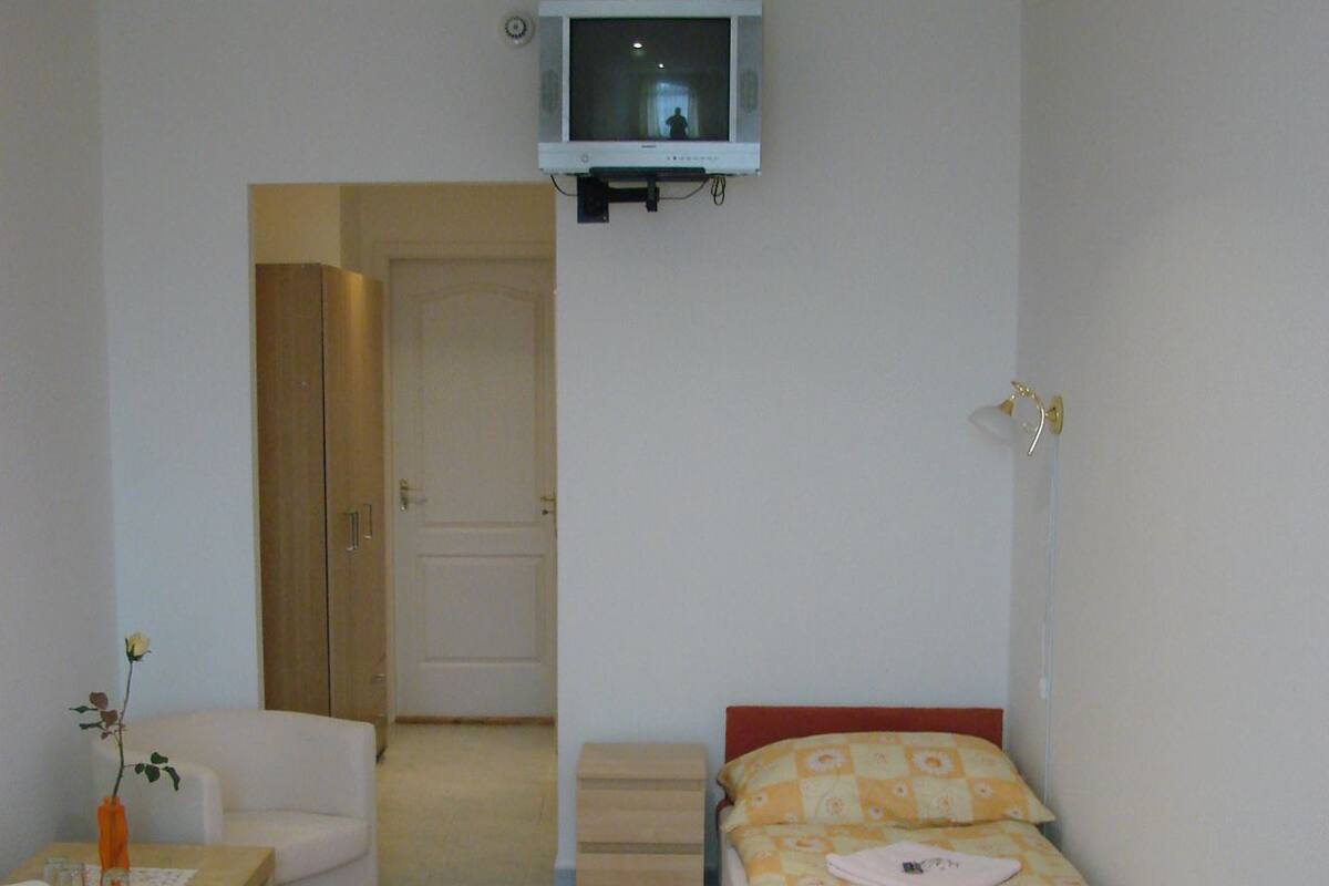 A small, well-lit bedroom with a single bed, a chair, a table with a vase, and a television mounted on the wall.