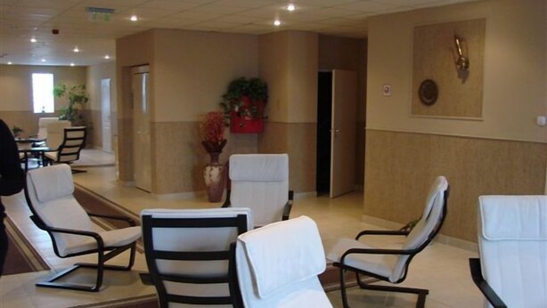A hotel lobby with several white cushioned chairs arranged in a semi-circle, a large vase with red flowers, and a wall-mounted clock. - Timon (Budapest)