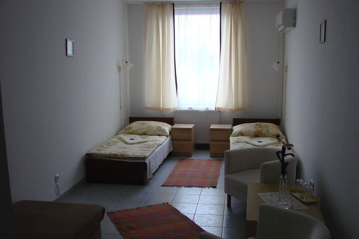 A small, well-lit room with two single beds, a small table, a chair, and a window with curtains.