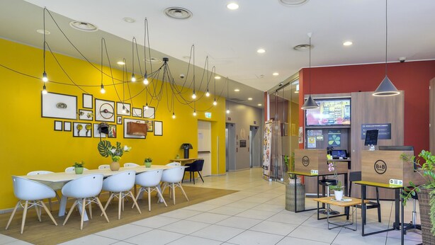 Un moderno vestíbulo de hotel con una pared amarilla, una recepción y una zona de estar con sillas y mesas.