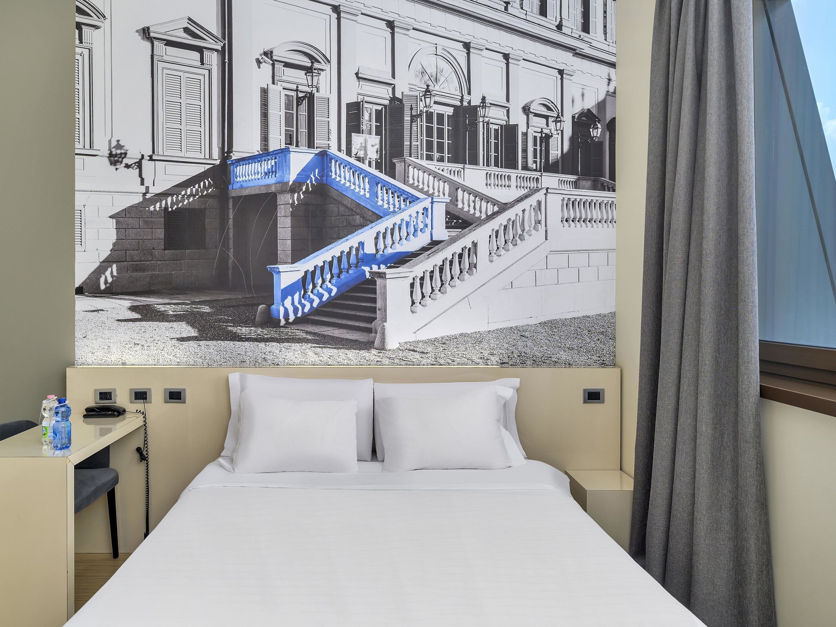 A hotel room with a bed, a nightstand, a lamp, and a view of a building with a blue staircase.