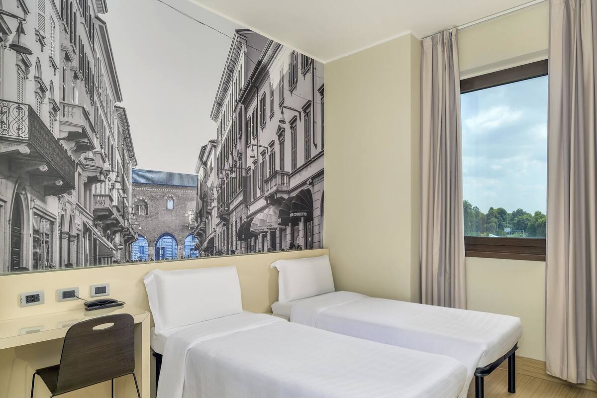 A hotel room with two beds, a desk, a chair, and a large window with curtains.