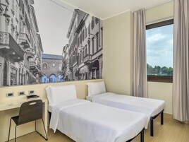 Quarto de hotel com duas camas, uma escrivaninha, uma cadeira e uma janela grande com cortinas.