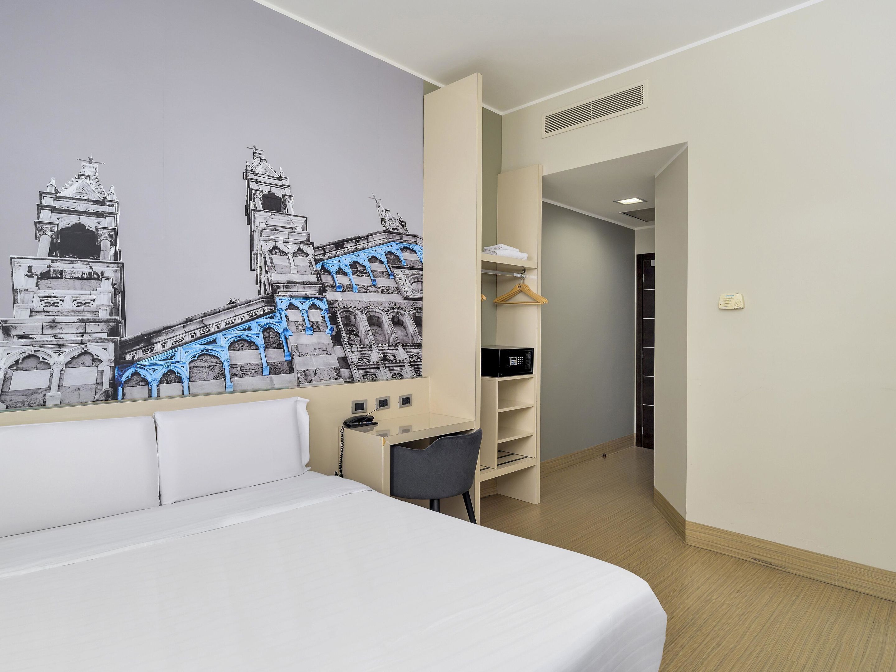 A hotel room with a bed, a desk, a chair, a wardrobe, and a wall mural of a historical building.