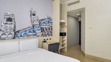 A hotel room with a bed, a desk, a chair, a wardrobe, and a wall mural of a historical building.