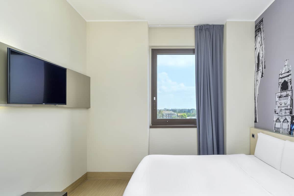 A hotel room with a bed, a flat-screen TV mounted on the wall, a window with a view of buildings, and a curtain.