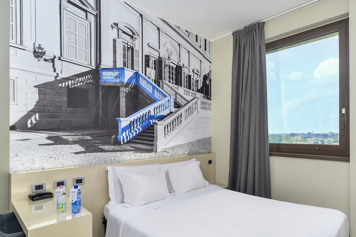 A hotel room with a bed, a desk with water bottles, a view of a cityscape through a window, and a mural of a staircase.