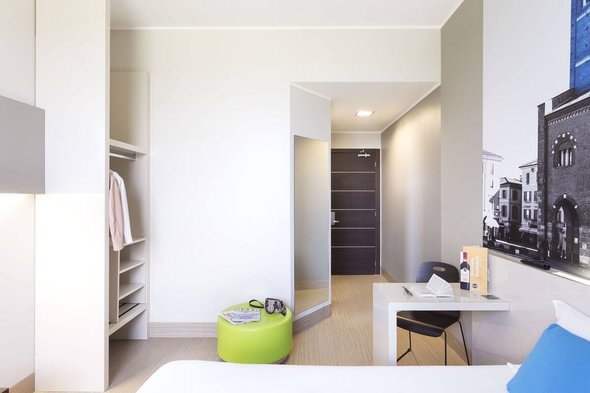 A modern hotel room with a walk-in closet, a green ottoman, a desk with a chair, and a cityscape view.