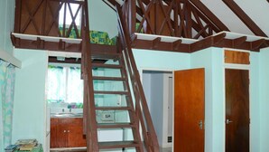 A room with a wooden staircase leading to a loft area, a kitchenette with a sink and cabinets, and two wooden doors. - Gina's Garden Lodges (Aitutaki)