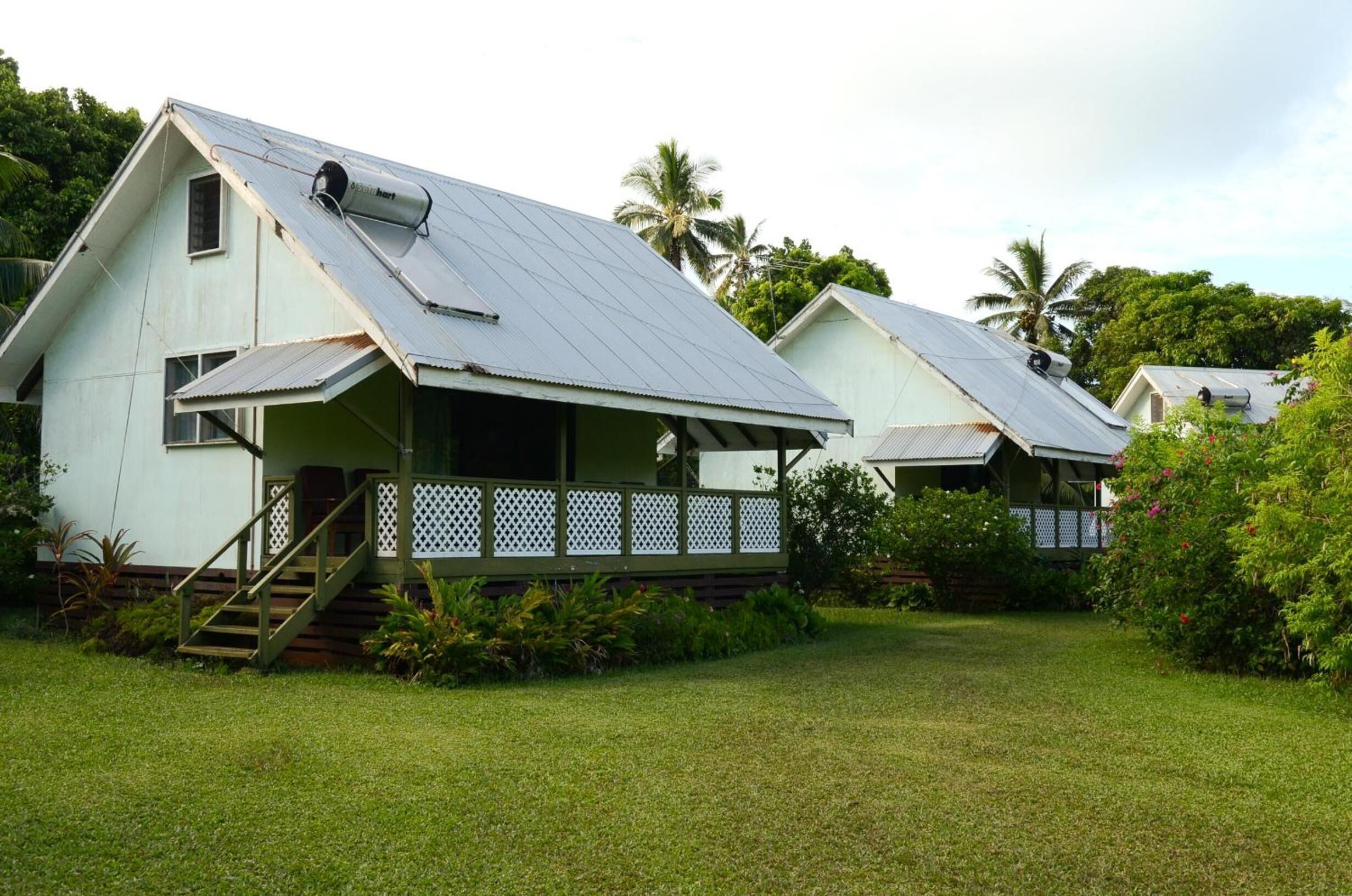 A row of white, single-story houses with metal roofs, surrounded by lush greenery and palm trees.