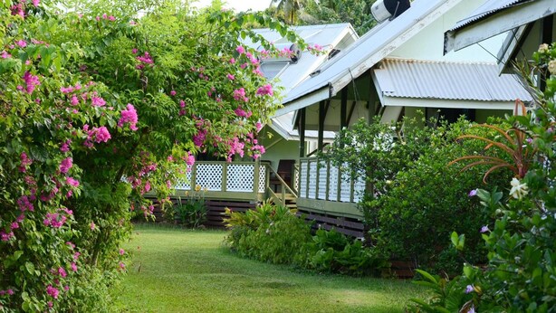 A house with a porch, surrounded by lush greenery and flowering plants. - Gina's Garden Lodges (Aitutaki)