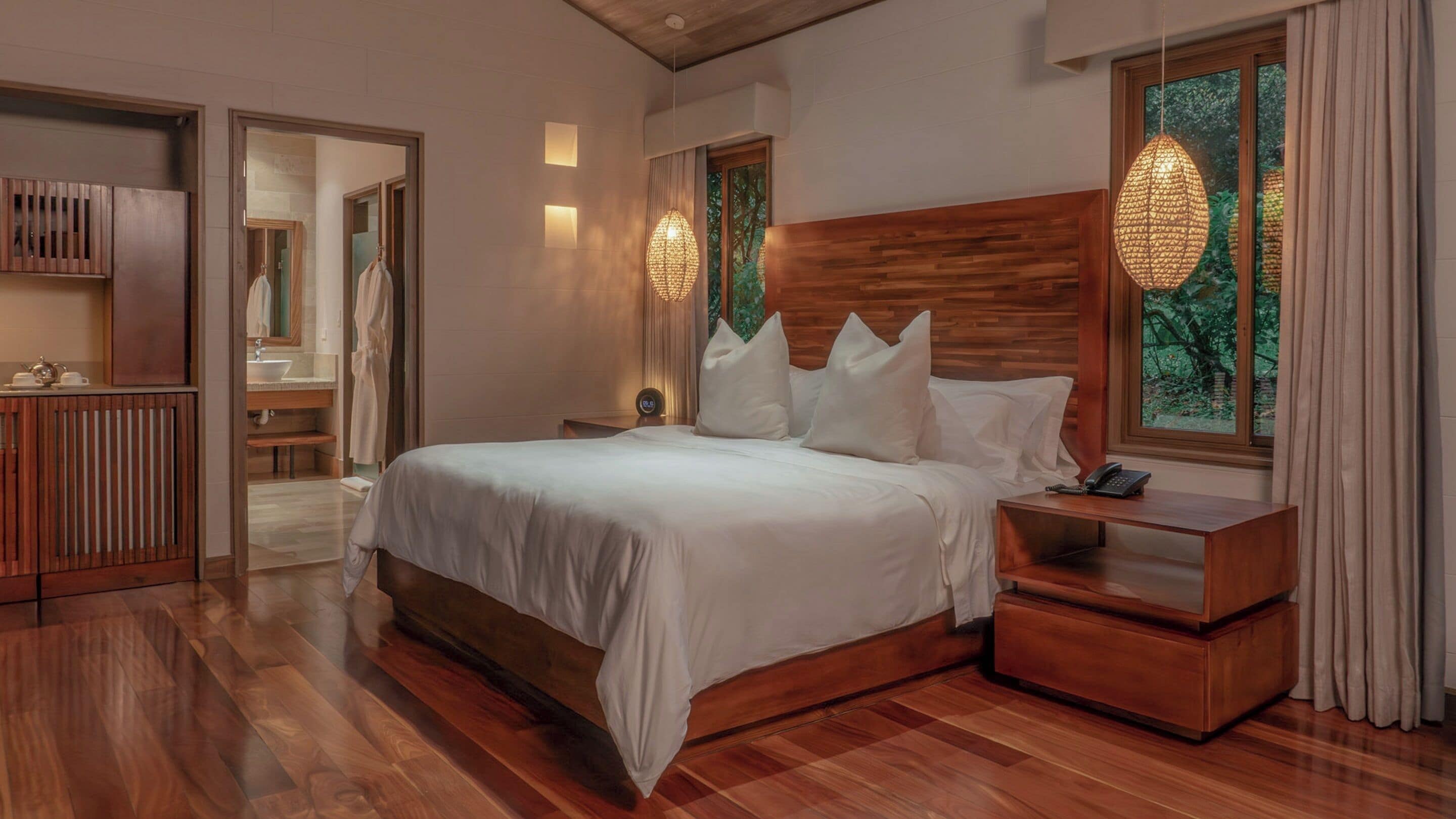 A bedroom with a wooden headboard, a large bed with white linens, a bedside table with a phone, and a window with curtains.