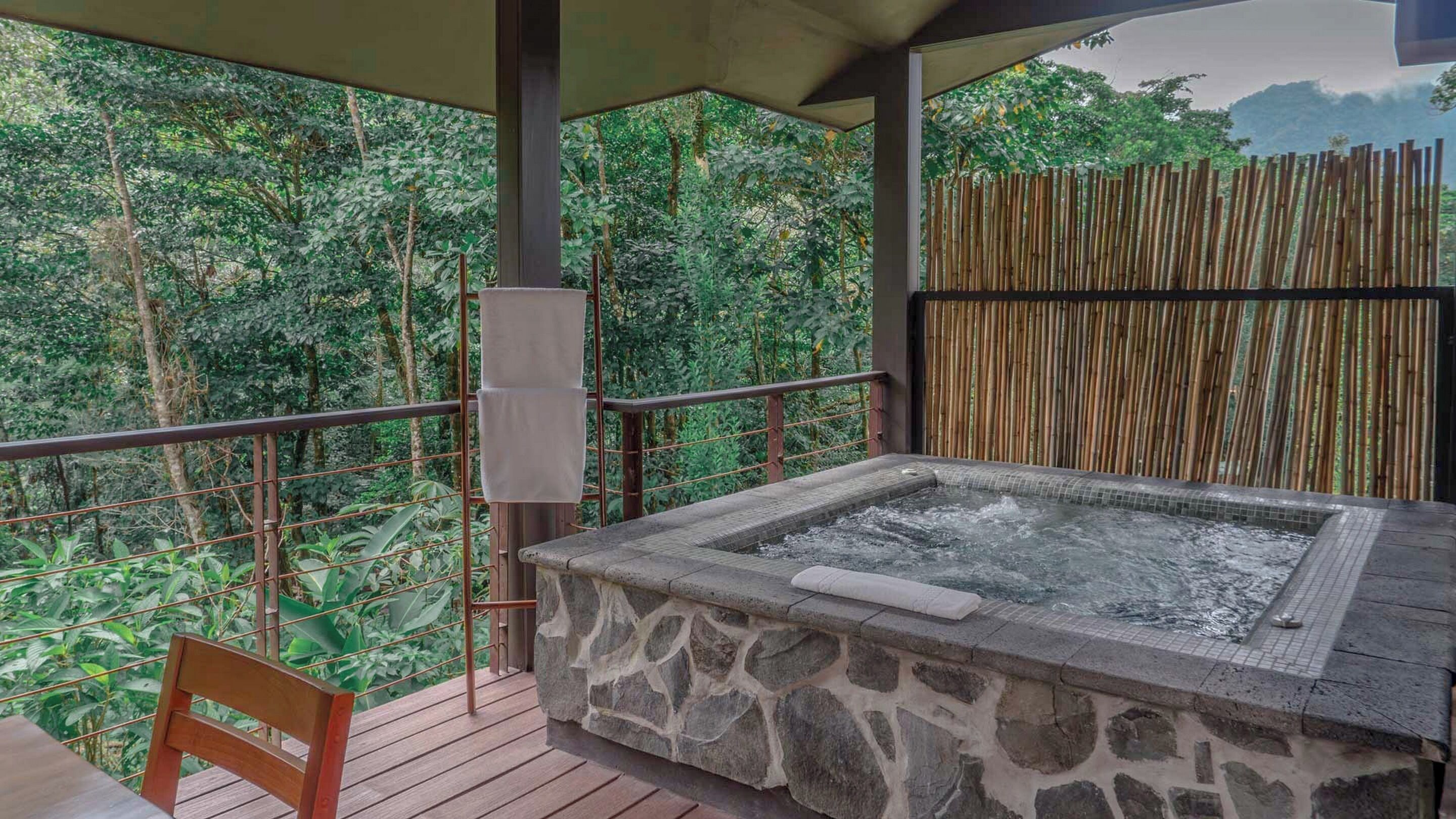 A hot tub on a wooden deck with a stone surround, surrounded by lush greenery and bamboo fencing.