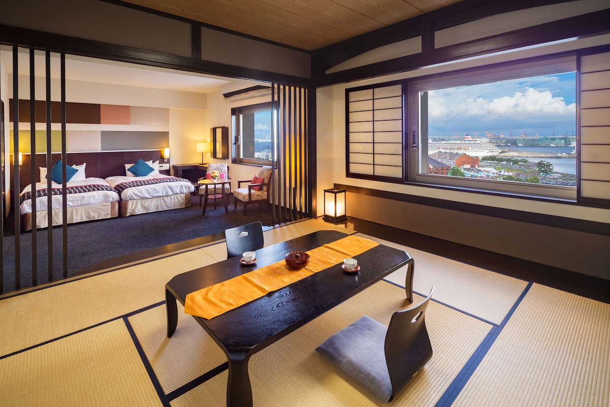 A traditional Japanese-style hotel room with tatami flooring, a low table with a yellow mat, and a view of a port with ships and cranes.