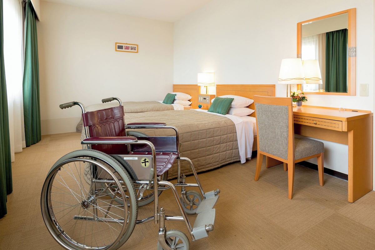 A hotel room with a wheelchair, two beds, a desk, and a mirror.