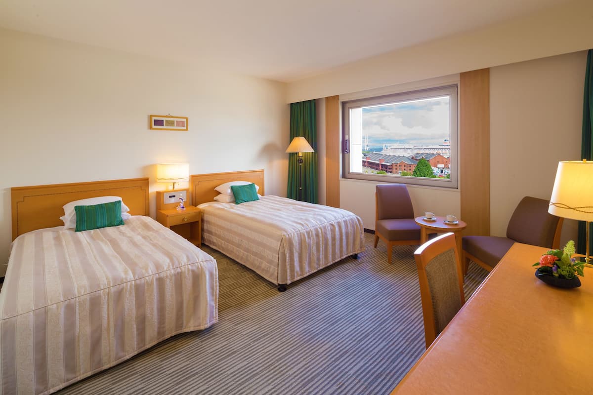 A hotel room with two beds, a desk, a chair, a small table, and a window with a view of buildings.