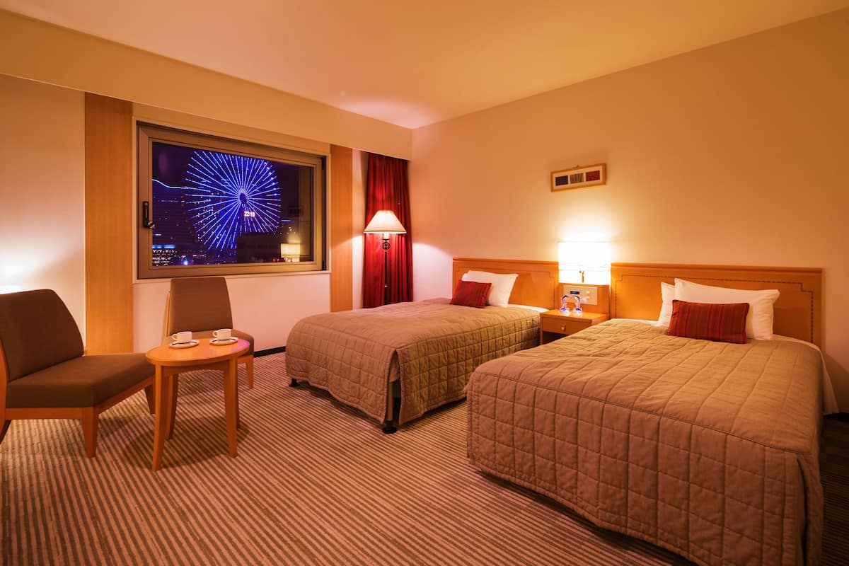A hotel room with two beds, a desk, a chair, a TV displaying a cityscape, and a lamp.