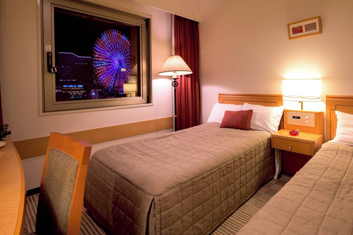 A hotel room with two beds, a nightstand, a lamp, a window with a view of a Ferris wheel, and a wall clock.